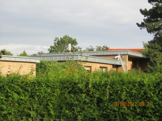 Gelbliches Backsteinhaus mit flachem, grauem Dach, vor einer grünen Hecke. Ein Mann steht auf einer Leiter am Haus. Im Vordergrund das Datum 18/09/2012 09:21.