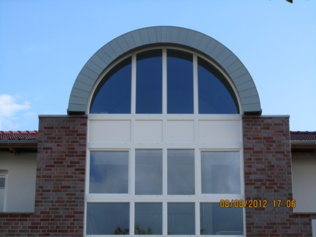 Großes, halbrundes Fenster mit blaugrauem Rahmen. Darunter rechteckige Fenster. Teil einer Ziegelwand.